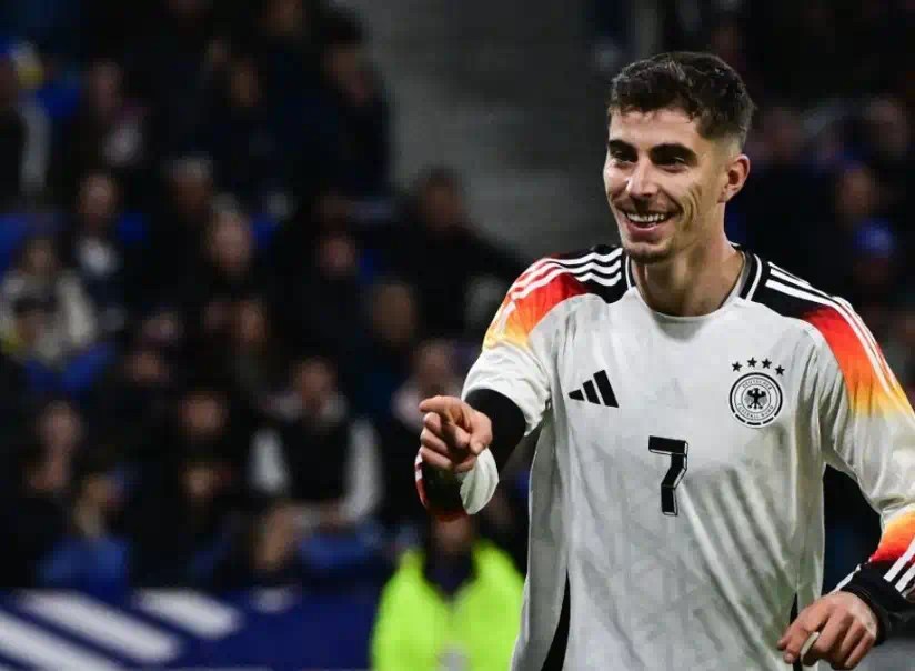 凱·哈弗茨(Kai Havertz)身穿德國國家隊白色球衣在比賽中面帶笑容並指向前方,背景為模糊的觀眾席與場上球員