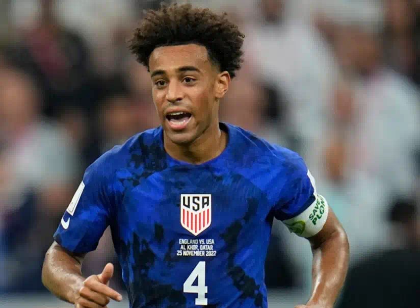 美國足球員泰勒·阿當斯(Tyler Adams)身穿藍色美國國家隊4號球衣,在比賽中向前跑動並專注觀察場上局勢,背景為模糊的觀眾看台。